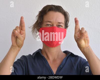 La femme caucasienne porte un masque de visage fait maison et croise ses doigts pour la bonne chance pendant la pandémie du virus corona 2019-20. Banque D'Images