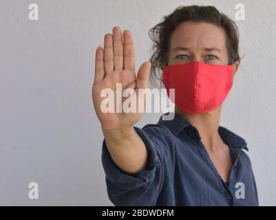 La femme caucasienne porte un masque de visage fait maison et fait le signe d'arrêt de paume de la main pendant la pandémie du virus corona 2019-20 - rester loin. Banque D'Images