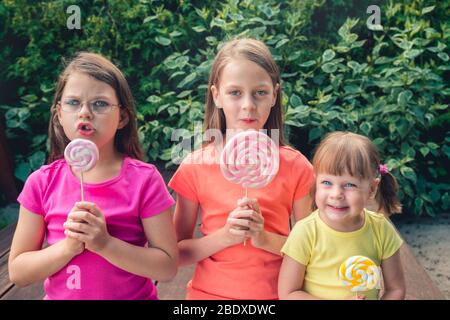 Trois petites filles drôles dans des tee-shirts colorés avec de grandes sucettes - sur la terrasse du jardin Banque D'Images