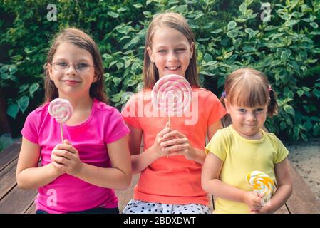 Trois petites filles drôles dans des tee-shirts colorés avec de grandes sucettes - sur la terrasse du jardin Banque D'Images