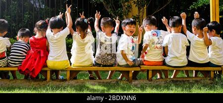 De jeunes enfants asiatiques heureux assis sur le banc de la maternelle un jour ensoleillé, à l'extérieur. Garderie, aire de jeux, jeux Banque D'Images