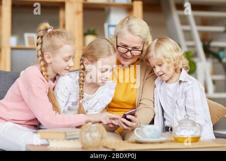 Femme senior dans les lunettes en tapant un message sur son téléphone mobile avec ses petits-enfants lorsqu'ils s'asseyent sur un canapé dans le salon Banque D'Images