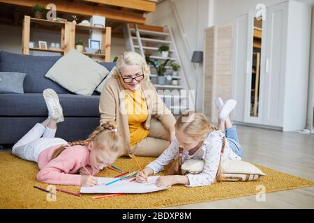 Deux petites filles dessinant avec des crayons sur le sol avec leur grand-mère assis près d'eux dans le salon Banque D'Images