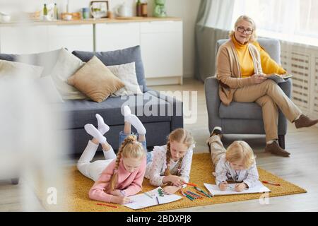 Groupe d'enfants qui dessinant des photos ensemble sur le sol avec grand-mère lisant un livre en arrière-plan Banque D'Images