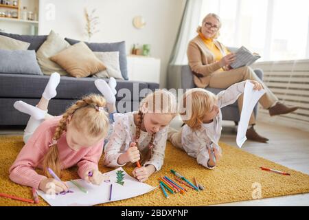 Les petites filles et les garçons qui se trouvent sur le sol et qui dessinant avec des crayons de couleur avec leur grand-mère lisent un livre dans un fauteuil en arrière-plan Banque D'Images