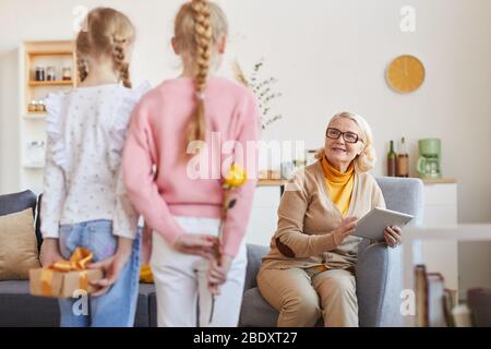 Grand-mère souriante assis sur un canapé et regardant ses petites-filles comment ils la félicitent avec des vacances Banque D'Images