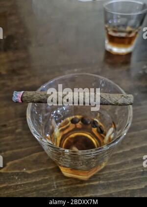 Gros plan sur un demi-verre de whisky au-dessus du bar en bois avec un cigare éclairé en plus, pourquoi fumer et l'alcoolisme est mauvais, partager le whisky scotch Banque D'Images