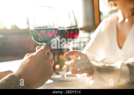 Les mains de deux jeunes dates amores s'inclinent avec des verres de vin rouge tout en étant assis par table servie dans un restaurant luxueux Banque D'Images