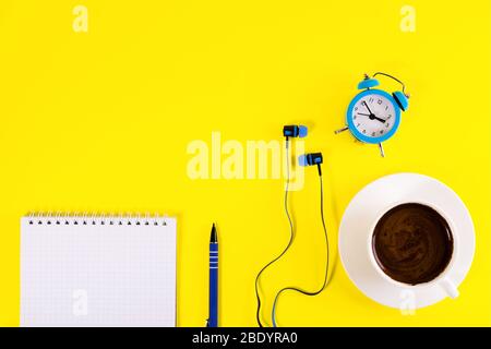 Le réveil bleu, les écouteurs bleus, la tasse de café et le carnet de notes avec le stylo bleu, se trouve sur fond jaune vif.étude et concept d'apprentissage. Espace de copie. Banque D'Images