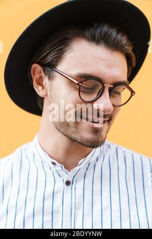 Image du jeune homme heureux portant des lunettes et un chapeau noir souriant et regardant vers le haut à l'espace publicitaire isolé sur fond jaune Banque D'Images