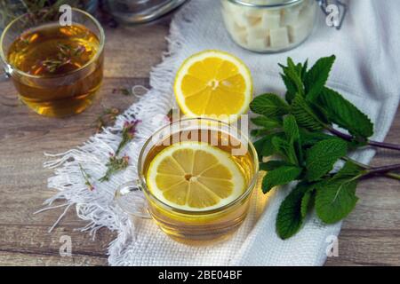 Tasse transparente en verre avec support à thé à la menthe sur un support en bois, à côté d'un bol à sucre avec des cubes de sucre, des feuilles de citron et de menthe tranchées Banque D'Images
