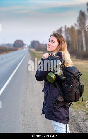 Une fille avec un masque à gaz noir sur son épaule se tient sur le bord d'une autoroute de banlieue. La fille essaie d'arrêter de passer des voitures afin de quitter la c Banque D'Images