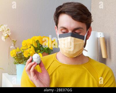 Homme caucasien tenant un oeuf de pâques avec un masque de visage pendant les heures de pâques, le temps d'éclosion de coronavirus, avec la bruyère jaune et les fleurs en arrière-plan. Banque D'Images