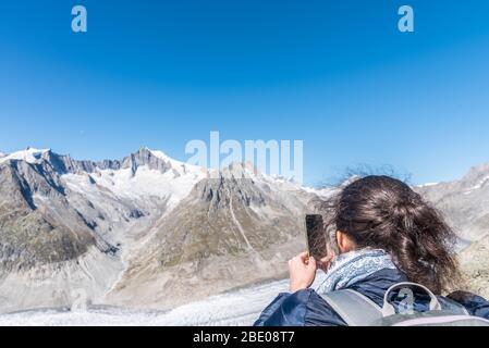 Vue arrière d'une femme photographiant le glacier d'Aletsch avec un smartphone du point de vue d'Eggishorn, les Alpes bernoises, le canton du Valais, la Suisse, l'Europe Banque D'Images