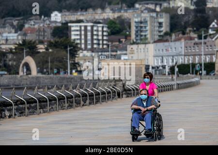 Une femme posant un homme en fauteuil roulant portant des masques de protection sur Marine Parade à Weston-super-Mare pendant le verrouillage pandémique de Coronavirus. Banque D'Images