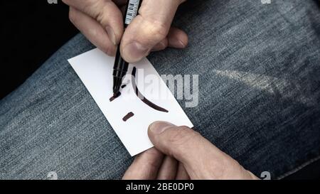 Écrire un émotiji sourire très triste sur la carte de papier blanc dans les mains sur le fond de texture de jeans. Triste humeur mauvais temps incolore Banque D'Images