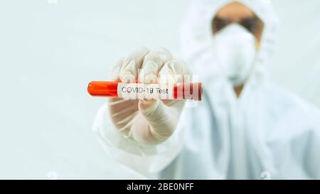 Médecin avec masque et costume blanc bioprotecteur avec bras étendu vers l'avant et sa main droite tenant un tube à essai avec échantillon de sang et étiquette Covid-19 dans Banque D'Images