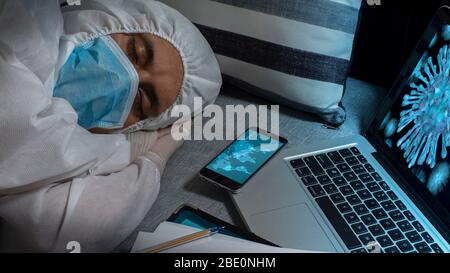 Médecin avec masque bleu et costume bioprotecteur dormant à côté de son ordinateur portable avec l'image du coronavirus sur l'écran et la carte de l'épidémie sur le sc Banque D'Images