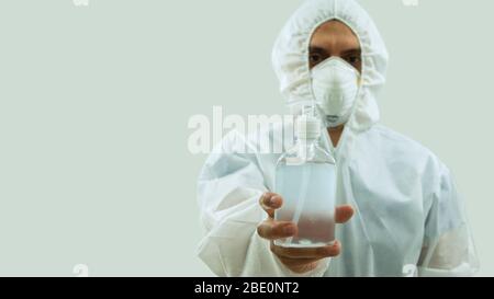 Médecin avec masque et costume blanc de bioprotection tenant avec sa main une bouteille d'alcool de gel au premier plan sur fond blanc Banque D'Images