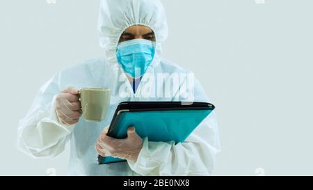 Médecin avec masque bleu et costume bioprotecteur lecture d'un dossier qu'il tient et une tasse de café verte dans sa main droite sur fond blanc Banque D'Images