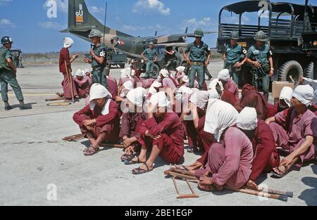 Prisonniers de Viet Cong s'asseoir sur la piste à la base aérienne de Tan Son Nhut, sous le regard vigilant de la police militaire du Sud Vietnam. Les prisonniers ont été amenés à la base aérienne dans le 6X6 camions dans l'arrière-plan et seront transportés par avion à Loc Ninh, Vietnam du Sud sur l'avion de transport C-123 pour l'échange de prisonniers entre les États-Unis et le Vietnam du Sud et Nord Vietnam/Viet Cong militaires. Banque D'Images