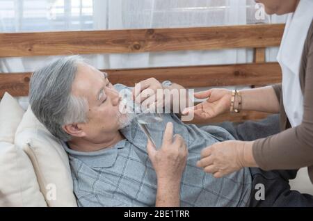 Homme asiatique principal prenant des médicaments et de l'eau potable tout en se trouvant sur le canapé. La vieille femme prend soin de son mari pendant que sa maladie à la maison. Banque D'Images