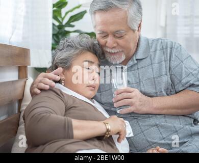 Femme asiatique senior prenant des médicaments et de l'eau potable tout en étant assise sur le canapé. L'homme âgé prend soin de sa femme pendant qu'elle est malade à la maison. Banque D'Images