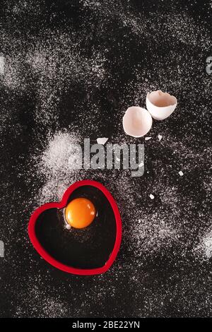 Image-clé faible d'un œuf biologique blanc cru sur fond noir saupoudrée de farine. Un œuf fissuré s'ouvre sur une forme en silicone rouge en forme de coeur. Cuisson Banque D'Images
