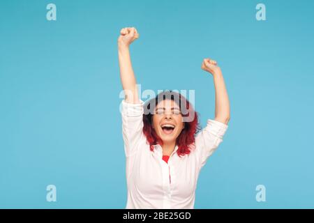 Je suis champion! Portrait d'une femme énergique et enthousiaste avec des cheveux rouges fantaisie dans une chemise qui se soulève les mains et criant de succès gagnant, vie heureuse. i Banque D'Images