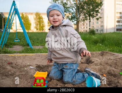 La Russie, Novodvinsk - juillet 2019. Juste un garçon brun de trois ans dans un chapeau bleu et un jean, dans une veste légère joue dans le bac à sable avec des jouets Banque D'Images