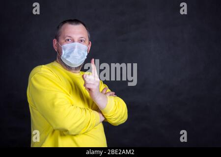 Homme d'âge moyen dans un masque jetable et une veste jaune maintient le pouce vers le haut Banque D'Images