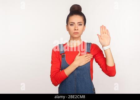 Je vous promets ! Portrait de la fille honnête digne de confiance avec un pain de cheveux dans des salopettes en denim soulevant la paume pour jurer l'allégeance, responsable et patriotique personne. ind Banque D'Images