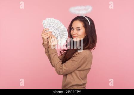 Portrait de jeune femme angélique joyeuse avec halo au-dessus de la tête tenant fan de billets en dollars, montrant de l'argent et se vantant de gagner de loterie, paradis riche Banque D'Images