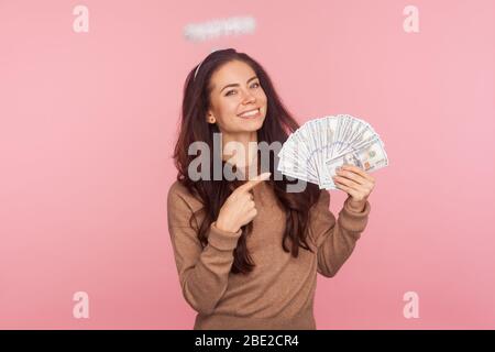 Portrait de la jeune femme angélique positive avec halo au-dessus de la tête pointant sur les billets en dollars, montrant de l'argent grand et se vantant de gagner de loterie, le paradis riche Banque D'Images