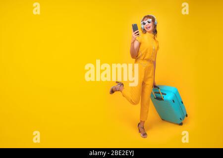 Jolie et drôle femme dans un style en tissu jaune décontracté tenant le téléphone cellulaire, portant des écouteurs et traînant des bagages bleus ou une valise dans le backroun de studio Banque D'Images