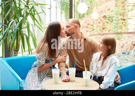 Père embrassant mère dans le café, assis à la table, leur fille les regarde étonnés. Environnement chaleureux, accueillant Banque D'Images