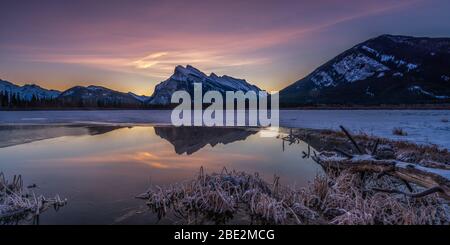 Le Mont Rundle a été recouvert de neige et un ciel coloré se reflète au lever du soleil dans les eaux fixes des lacs Vermillion, dans le parc national Banff, à Banff, en Alberta, au Canada