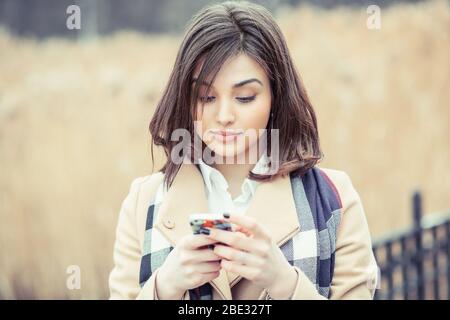 Portrait d'une belle femme avec des vêtements de printemps dactylographiant sur le smartphone dans un parc avec un fond jaune non focalisé Banque D'Images