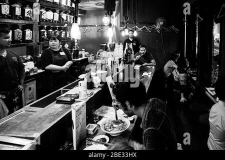Une serveuse rit derrière le comptoir du restaurant Jomon dans le quartier de Minato à Tokyo, au Japon. Un client au bar mange dans un bol. Un ventilateur de plafond tourne au-dessus du bar. Des bouteilles de saké arrondies s'alignent sur des étagères en bois le long du mur. Banque D'Images