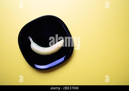une image moderne couleur pastel d'une banane une plaque carrée noire décorative avec fond jaune pastel, plat, vue sur le dessus Banque D'Images