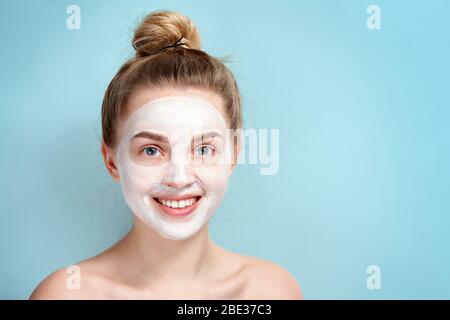 Jeune fille dans un masque cosmétique de visage blanc souriant. Soin de la peau. Blonde femme souriant sur fond bleu. Masque hydratant. Belle face de jeune adul Banque D'Images