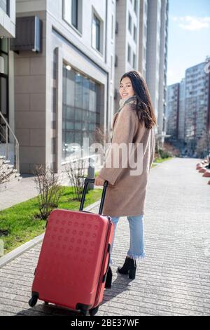 Jeune femme aux cheveux sombres, blouse beige portant une valise rouge Banque D'Images