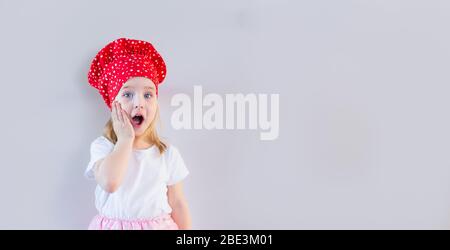 Petite fille blonde souriante dans le chapeau de cuisinier rouge restant sur fond gris neutre et tenant sa joue, surparée, choquée. Espace pour le texte. Banque D'Images