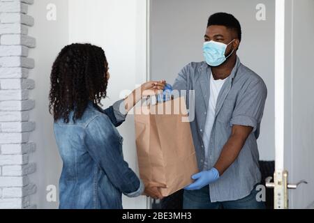 Un gars de messagerie noir dans un masque médical livrant une commande d'épicerie à une femme Banque D'Images