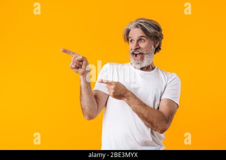 Image d'un homme barbu souriant aux cheveux gris, isolé sur fond de mur jaune pointant. Banque D'Images