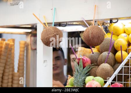 Avec la paille de noix de coco ouverte et d'autres fruits comme l'ananas orange. Un colporteur prépare les jus. Deux jeunes vendeurs dans le dos. Banque D'Images