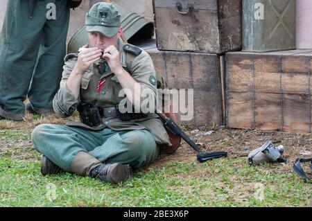 Un soldat assis sur le terrain, jouant une harmonica, vêtu d'un uniforme vert lors d'une réadoption de la seconde Guerre mondiale. Banque D'Images