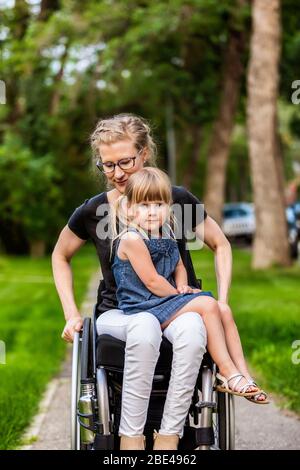 Une maman paraplégique tenant sa petite fille sur ses genoux tout en la prenant pour une promenade dans la rue dans son fauteuil roulant, un après-midi d'été chaud: Edmon... Banque D'Images