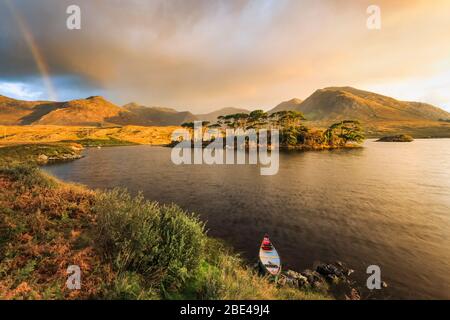 Canoë sur les rives d'un lac dans Connemara avec une île avec des arbres au lever du soleil avec des nuages de tempête et un arc-en-ciel au loin Banque D'Images