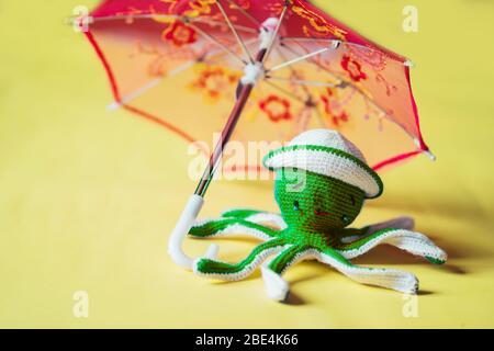 Mignonne poupée octope en crochet isolée sur fond jaune. Pieuvre verte et ludique portant un chapeau blanc assis sous un parapluie. Chil tricoté à la main Banque D'Images